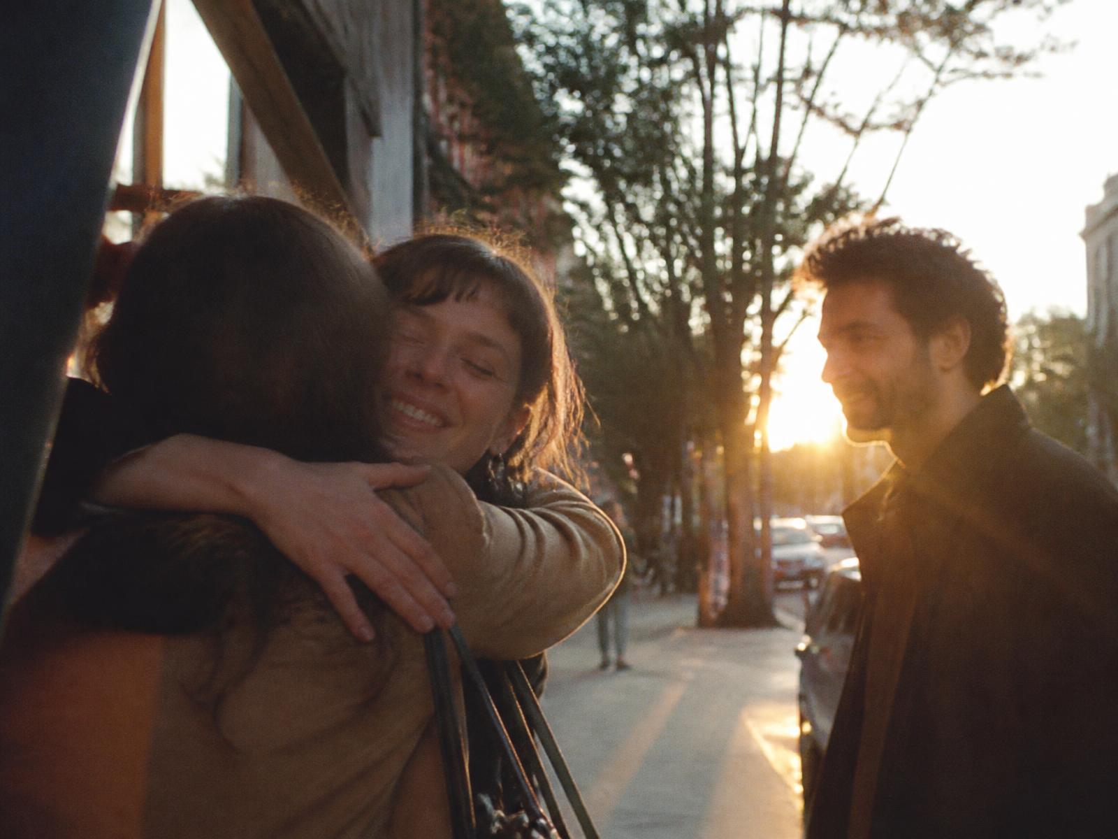 Two friends hugging goodbye on a sunlit street while a third waits nearby