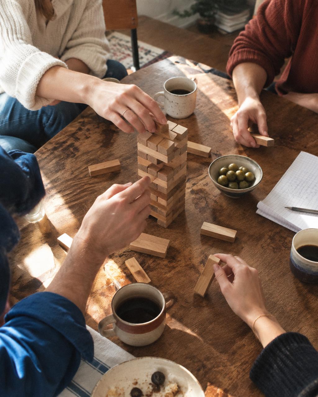 Hands around a wooden block-tower game on a sunlit table with coffee between friends